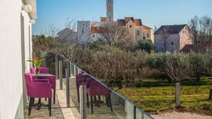A balcony with purple chairs and a table, overlooking a garden and buildings with a tower.
