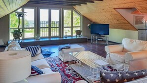 A spacious living room with a high ceiling, large windows, a television, and a cozy seating area with a patterned rug.