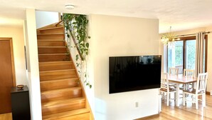 A modern living room with a wooden staircase, a flat-screen TV mounted on the wall, and a dining area with white chairs. - Sunny 3 bedroom cape with large deck, porch and kayaks. Pet friendly. (Oak Bluffs)