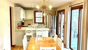 A dining area with a wooden table and white chairs, a kitchen with white cabinets, and a large window with curtains. - Sunny 3 bedroom cape with large deck, porch and kayaks. Pet friendly. (Oak Bluffs)