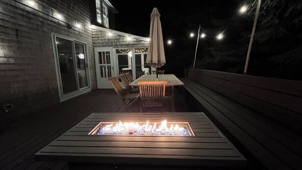 An outdoor patio with a fire pit, wooden deck, and a table with chairs. - Sunny 3 bedroom cape with large deck, porch and kayaks. Pet friendly. (Oak Bluffs)