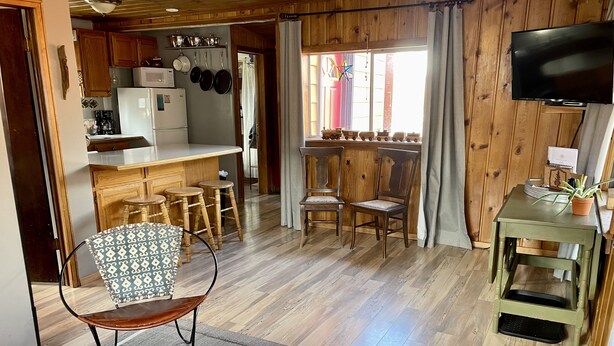 Una sala de estar acogedora con piso de madera, una mesa de centro, televisor y una zona de cocina con gabinetes de madera y un microondas.