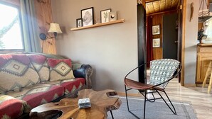Una sala de estar acogedora con una mesa de centro de madera, un sofá estampado y un sillón con estampado. Se observa una estantería con cuadros en la pared y una lámpara sobre una mesita auxiliar.