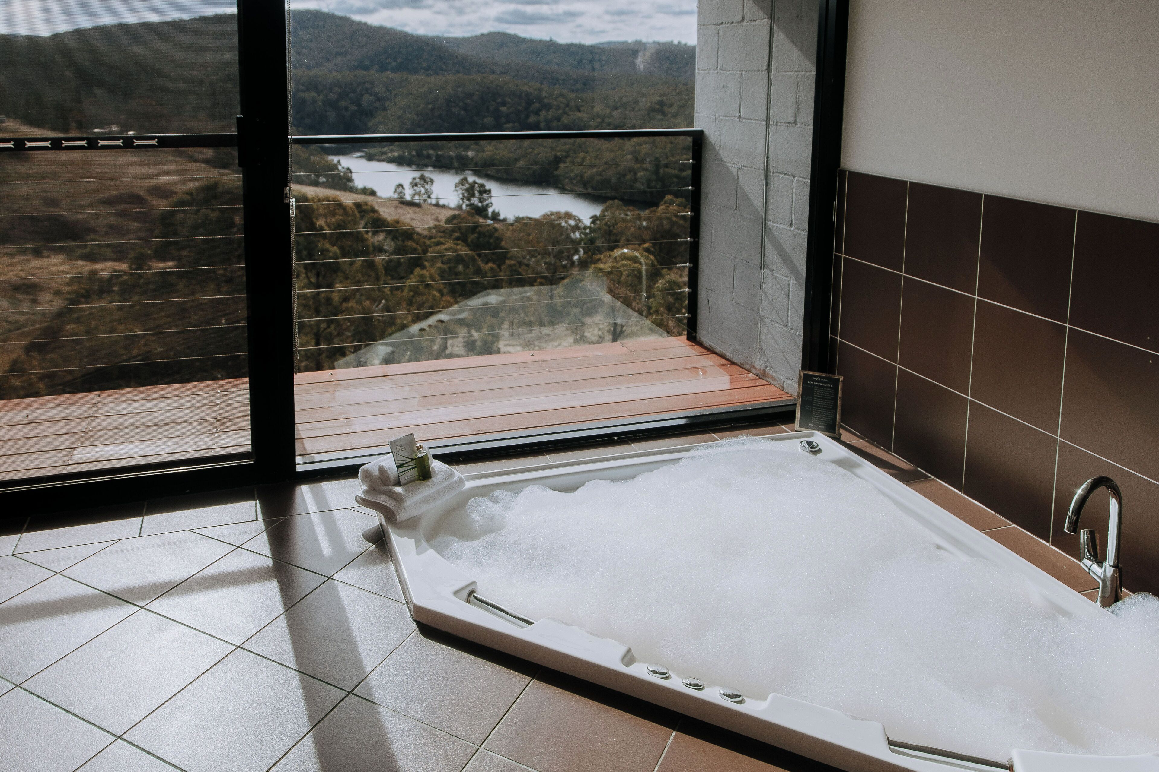 A modern bathroom with a large bathtub, a view of a river and forest through a glass door, and a tiled floor.