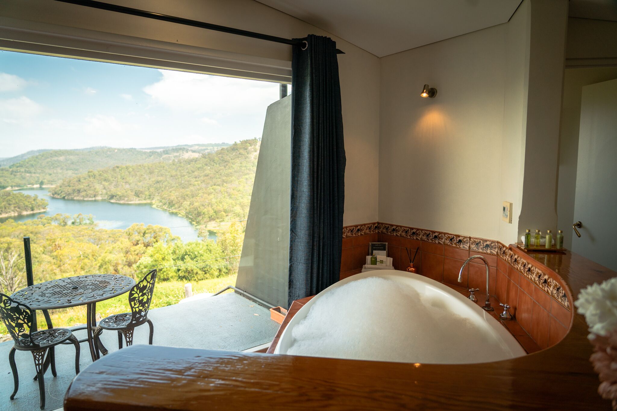 Un dormitorio con una bañera grande, una mesa de comedor con sillas y vistas a un lago y un bosque.