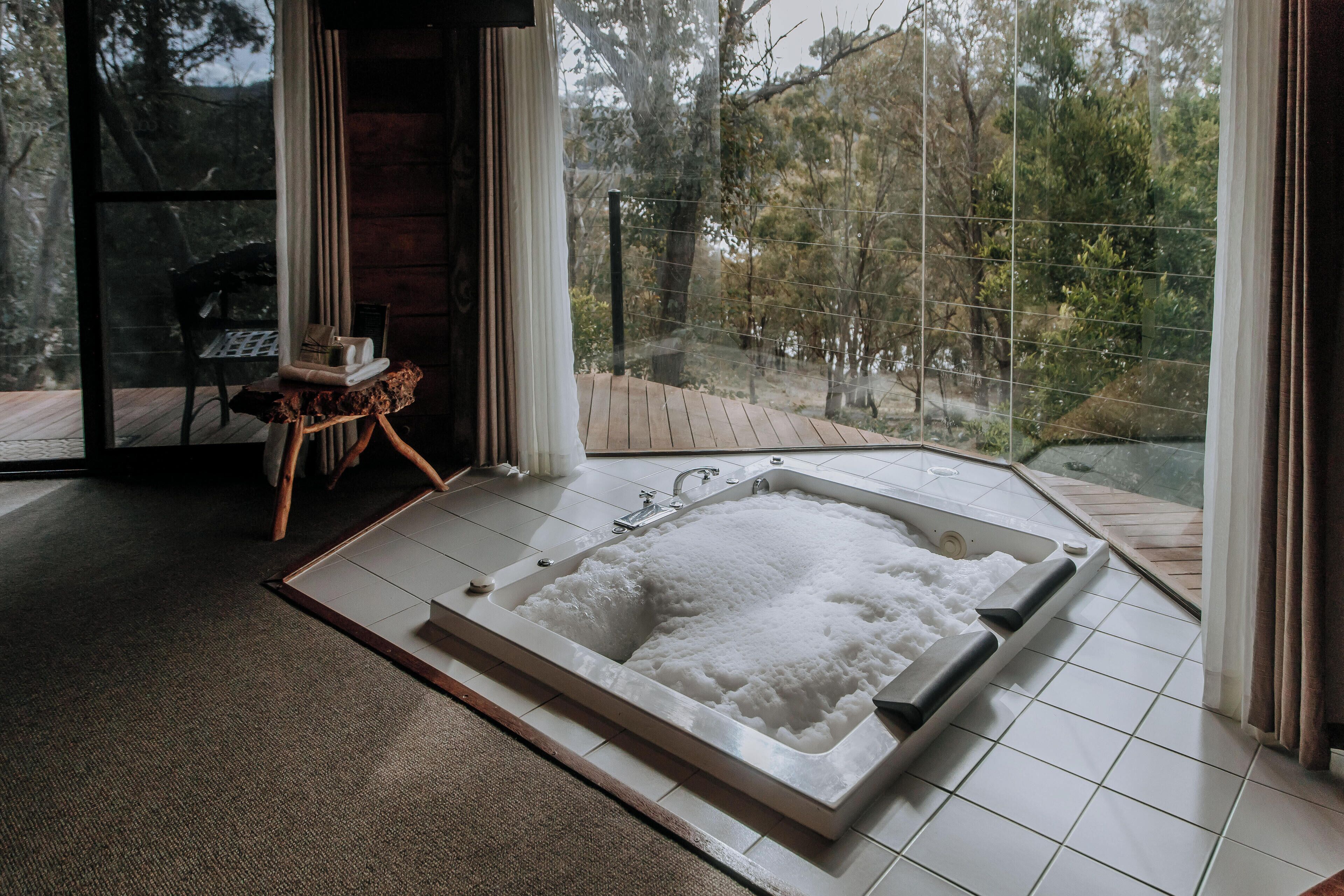 A modern bathroom with a large bathtub, a wooden side table, and a view of a forest through large windows.
