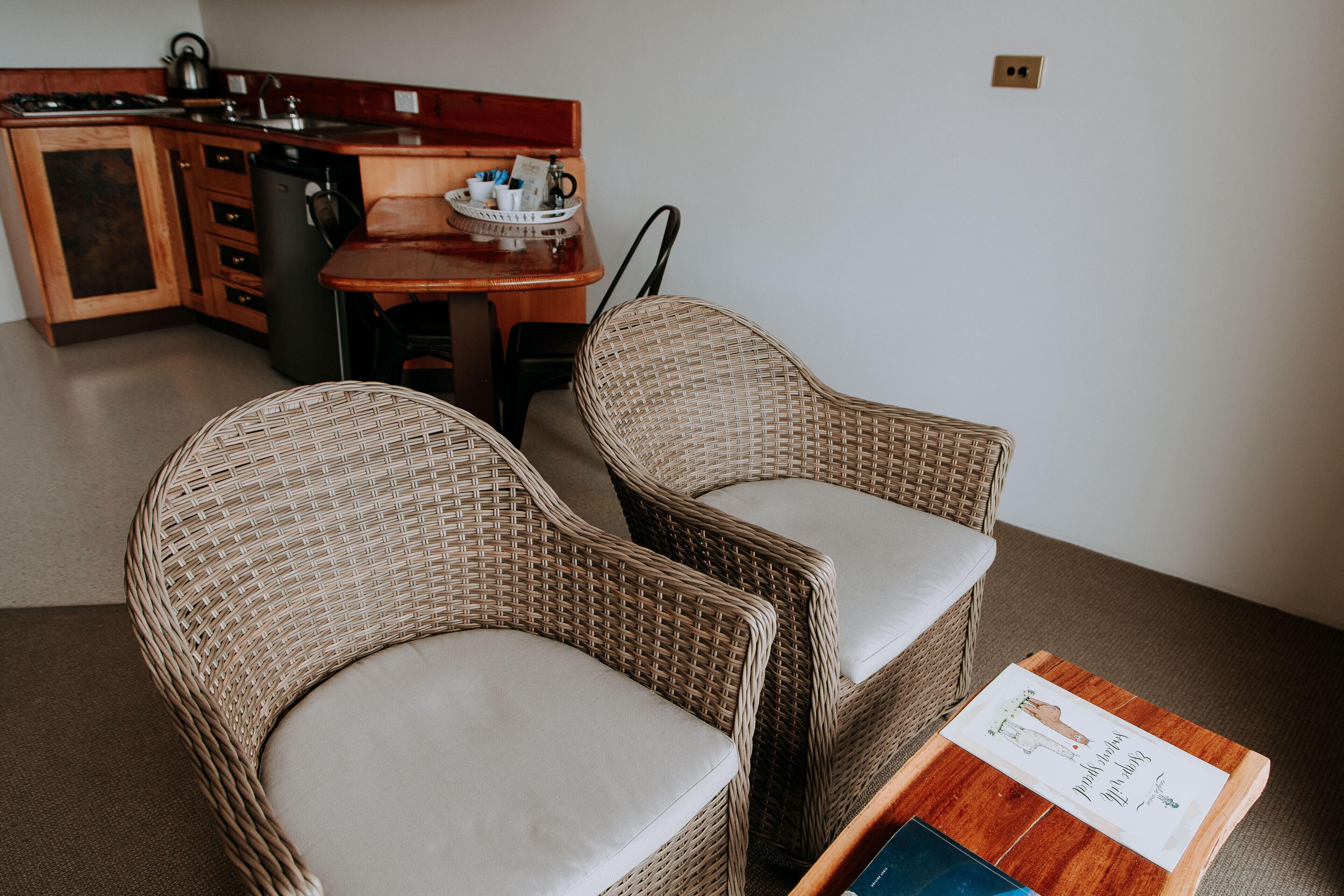 Une petite cuisine équipée d’une cuisinière, d’un évier et d’un petit réfrigérateur. On y trouve deux fauteuils en osier avec des coussins et une table en bois sur laquelle est posé un plateau contenant divers objets.