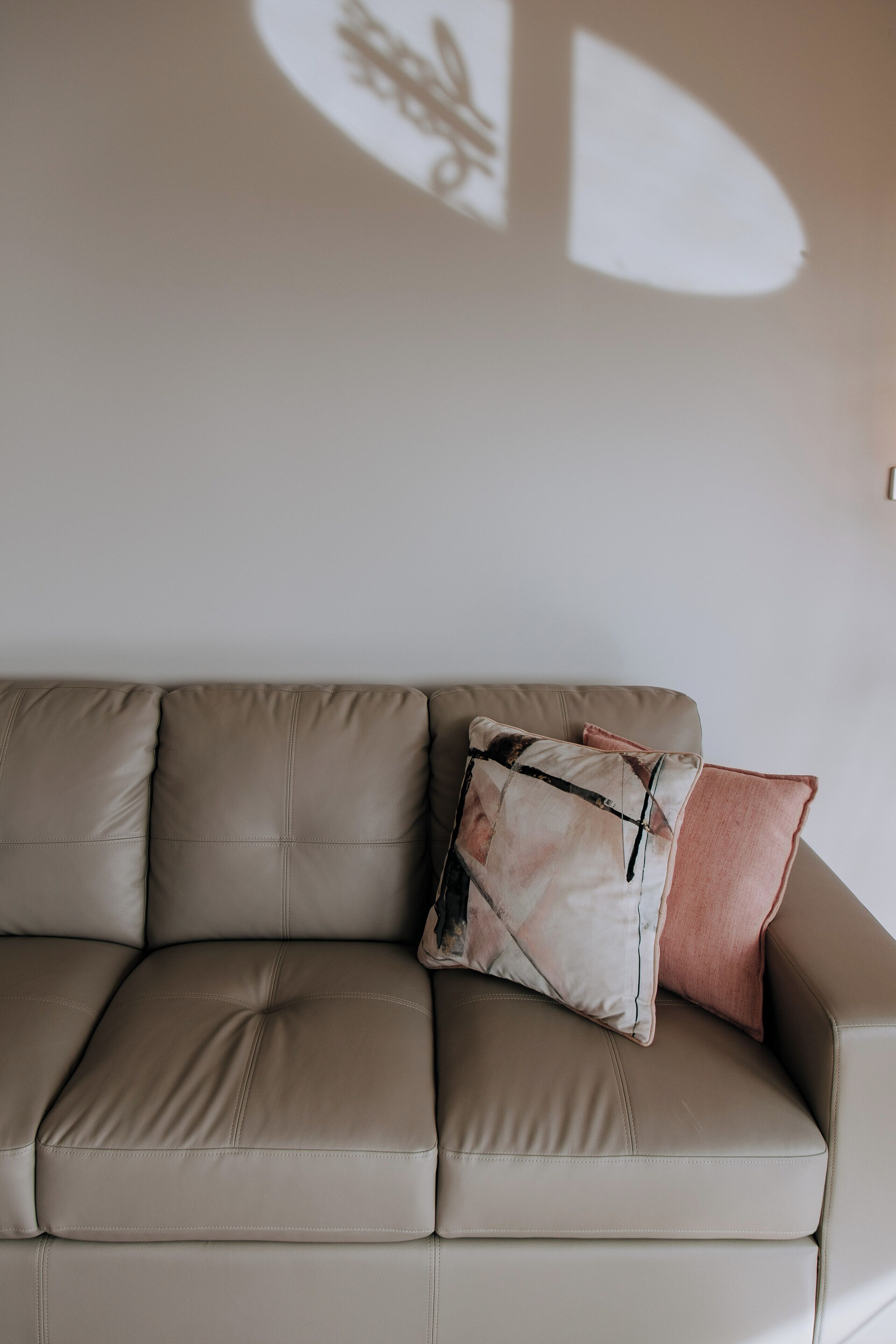 Un sofá de cuero color beige con dos almohadones, uno con estampado floral y otro de color liso, frente a una pared lisa.
