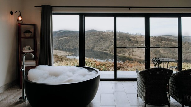 A modern bathroom with a large black bathtub filled with bubbles, a wooden shelf with toiletries, a wicker chair, and a large window offering a view of a mountainous landscape. - Eagle View Escape (Rydal)