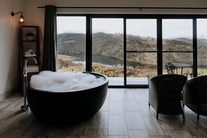 A modern bathroom with a large black bathtub filled with bubbles, a wooden shelf with toiletries, a wicker chair, and a large window offering a view of a mountainous landscape.