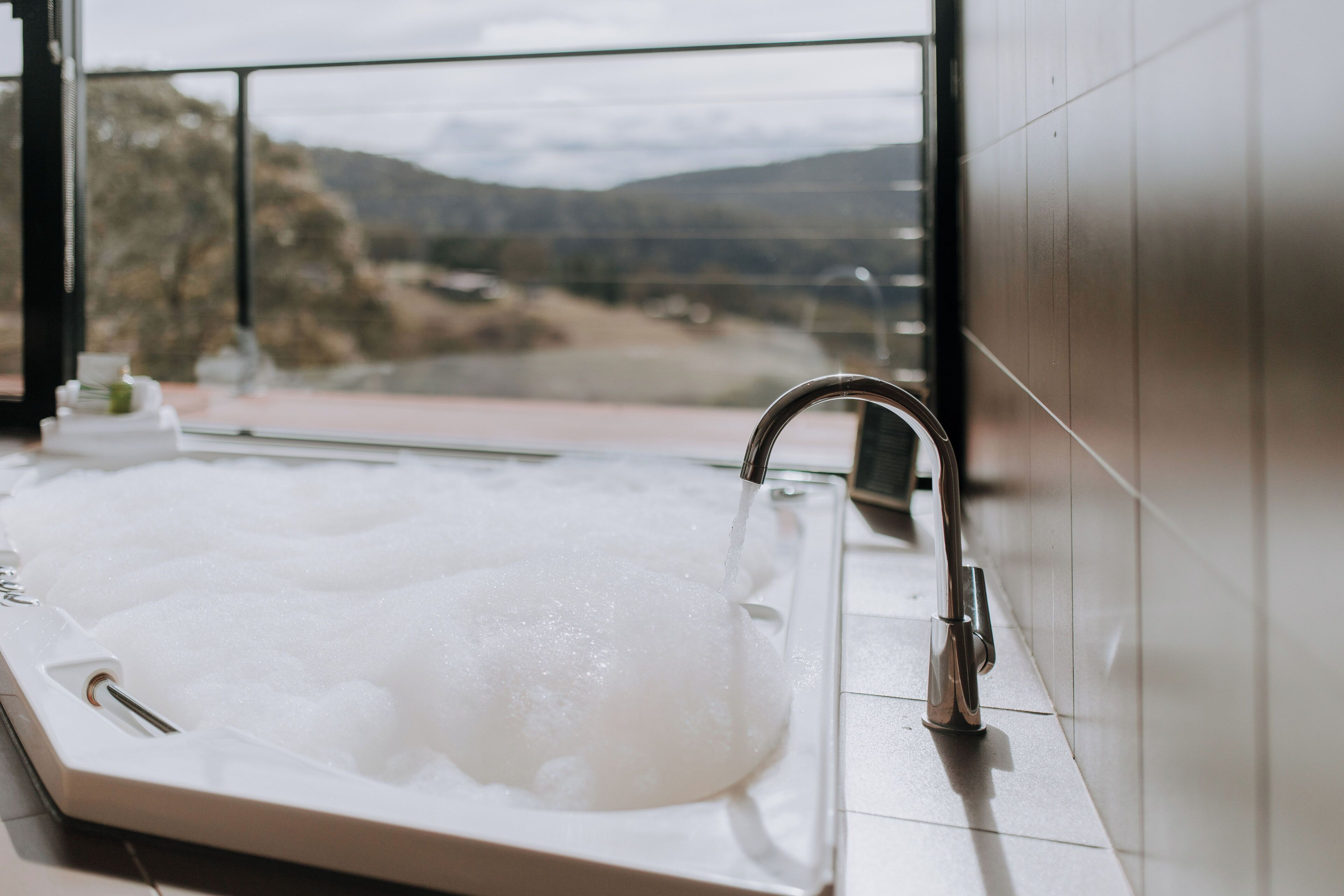 A modern bathroom with a large bathtub filled with bubbles, a faucet, and a view of a mountainous landscape through a large window.