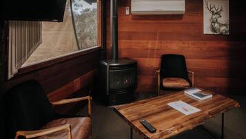 Une pièce chaleureuse avec un poêle à bois, des fauteuils en cuir, une table en bois sur laquelle reposent une télécommande et un livre, et un tableau représentant un cerf accroché au mur.