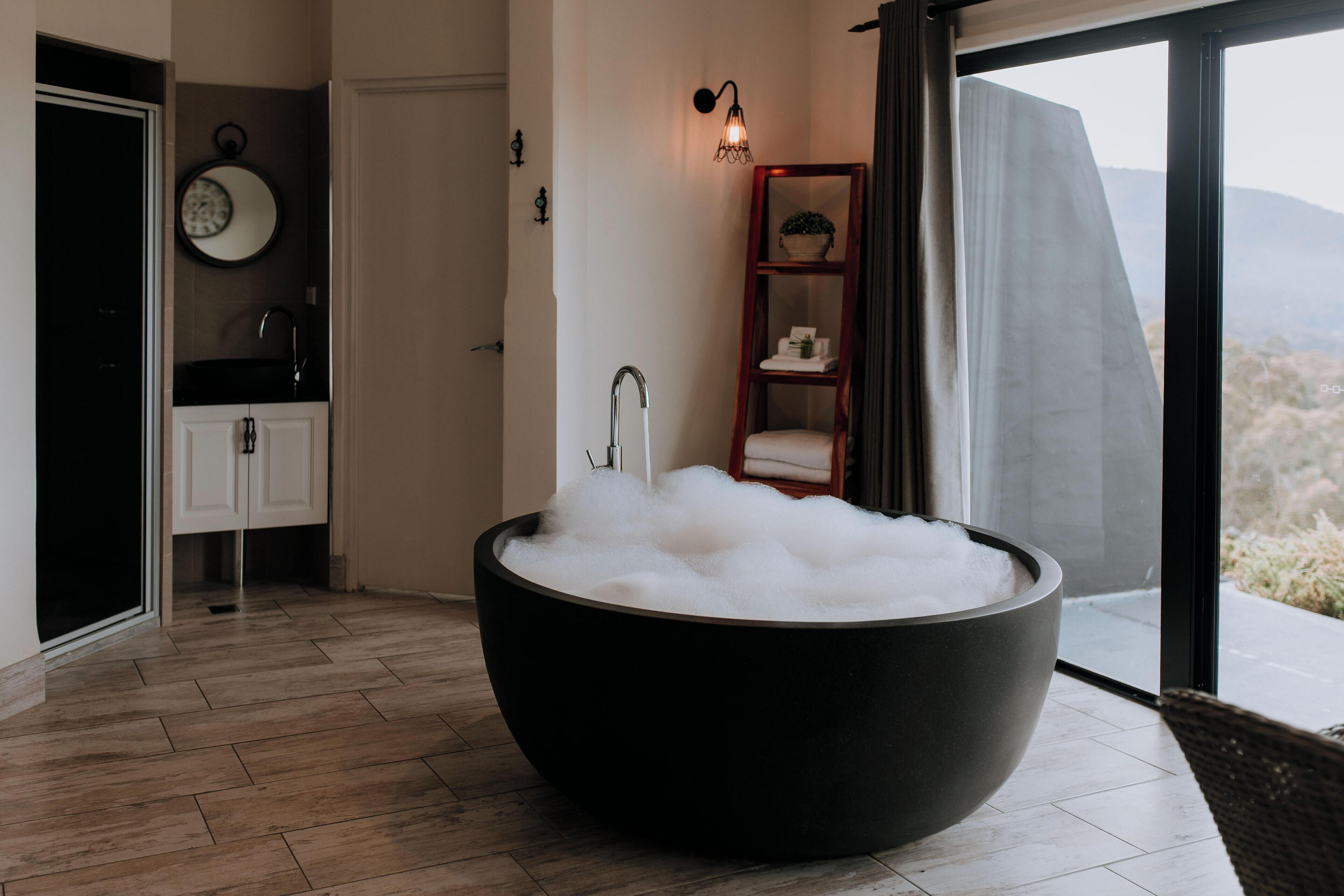 A modern bathroom with a large black bathtub filled with bubbles, a white sink, a wooden shelf with a plant, and a view of the desert through a large window.