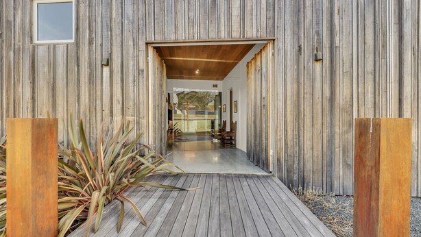 Fachada de madera con puerta de vidrio, una terraza de madera y una planta en maceta.