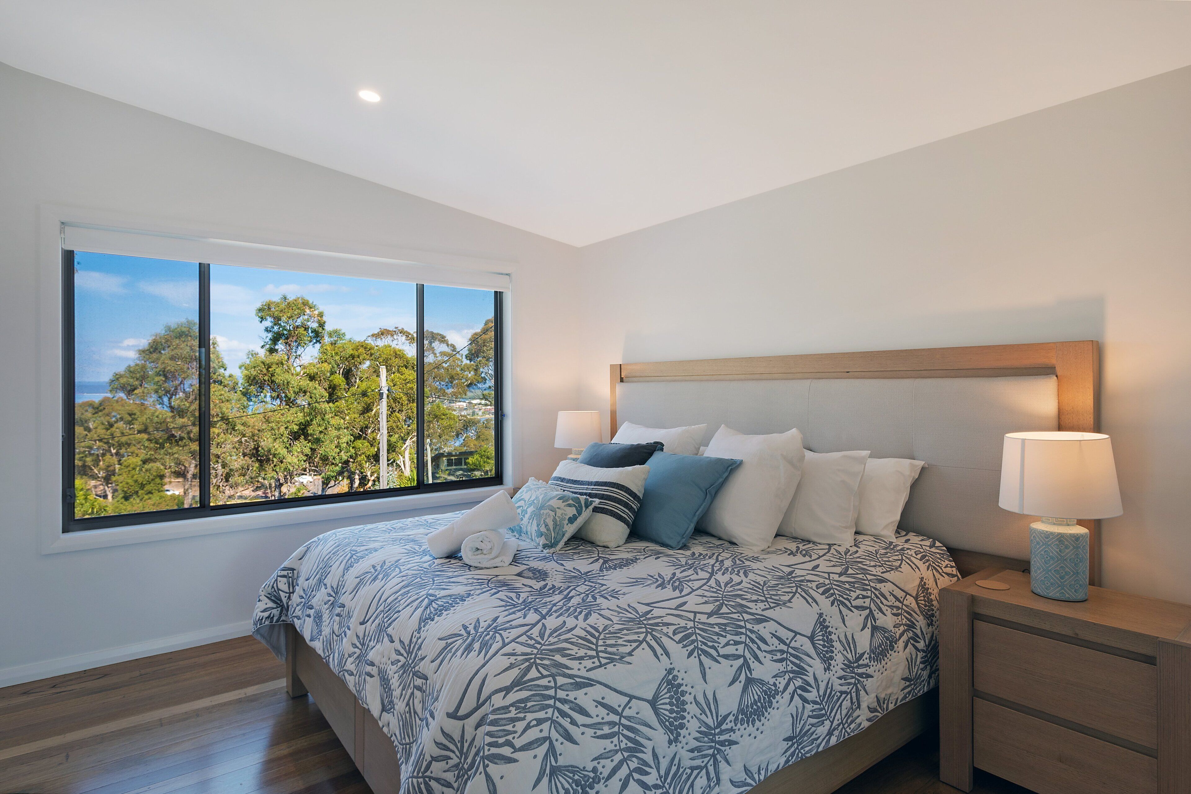 A bedroom with a large window, a bed with a floral bedspread, a nightstand with a lamp, and a view of trees outside.