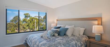 A bedroom with a large window, a bed with a floral bedspread, a nightstand with a lamp, and a view of trees outside.