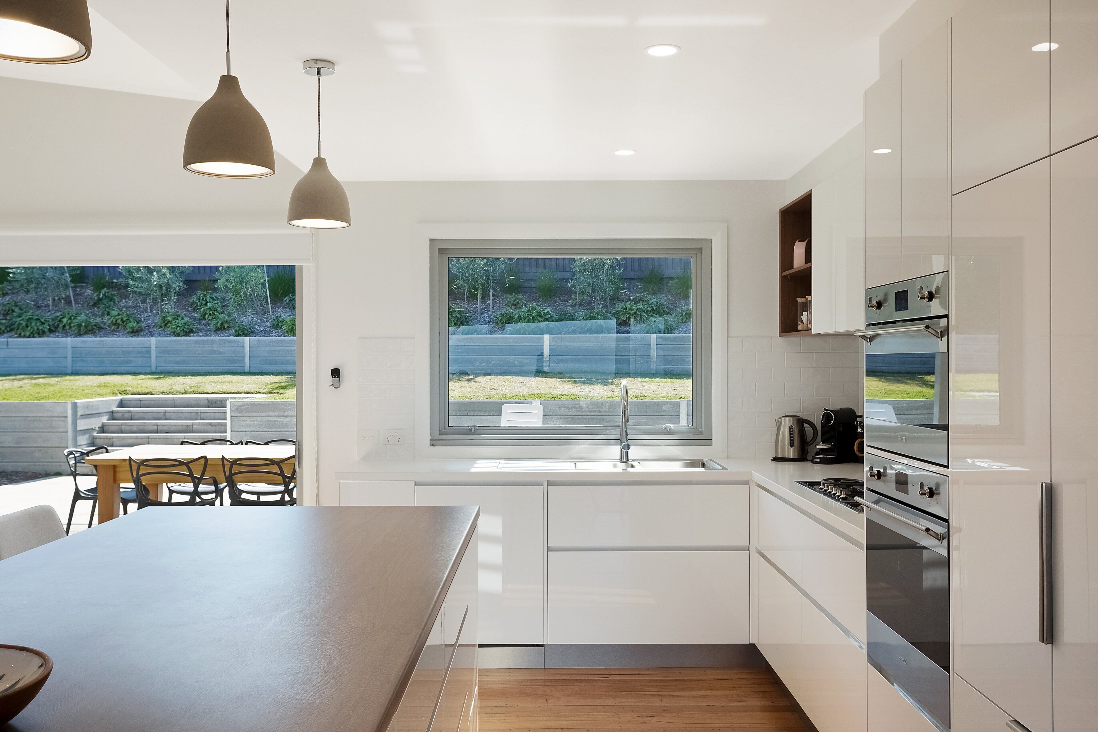 Une cuisine moderne avec des meubles blancs, une grande fenêtre et un coin repas comprenant une table et des chaises en bois.