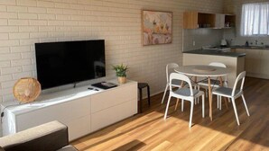 Un soggiorno moderno con una TV a schermo piatto, un piccolo mobiletto bianco, un tavolo da pranzo rotondo con sedie bianche e un angolo cottura sullo sfondo.