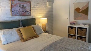 Una camera da letto con parete in mattoni bianchi, un letto con copripiumino a righe bianche e grigie, due cuscini, una lampada da comodino e un piccolo comodino bianco con libri e cesti.