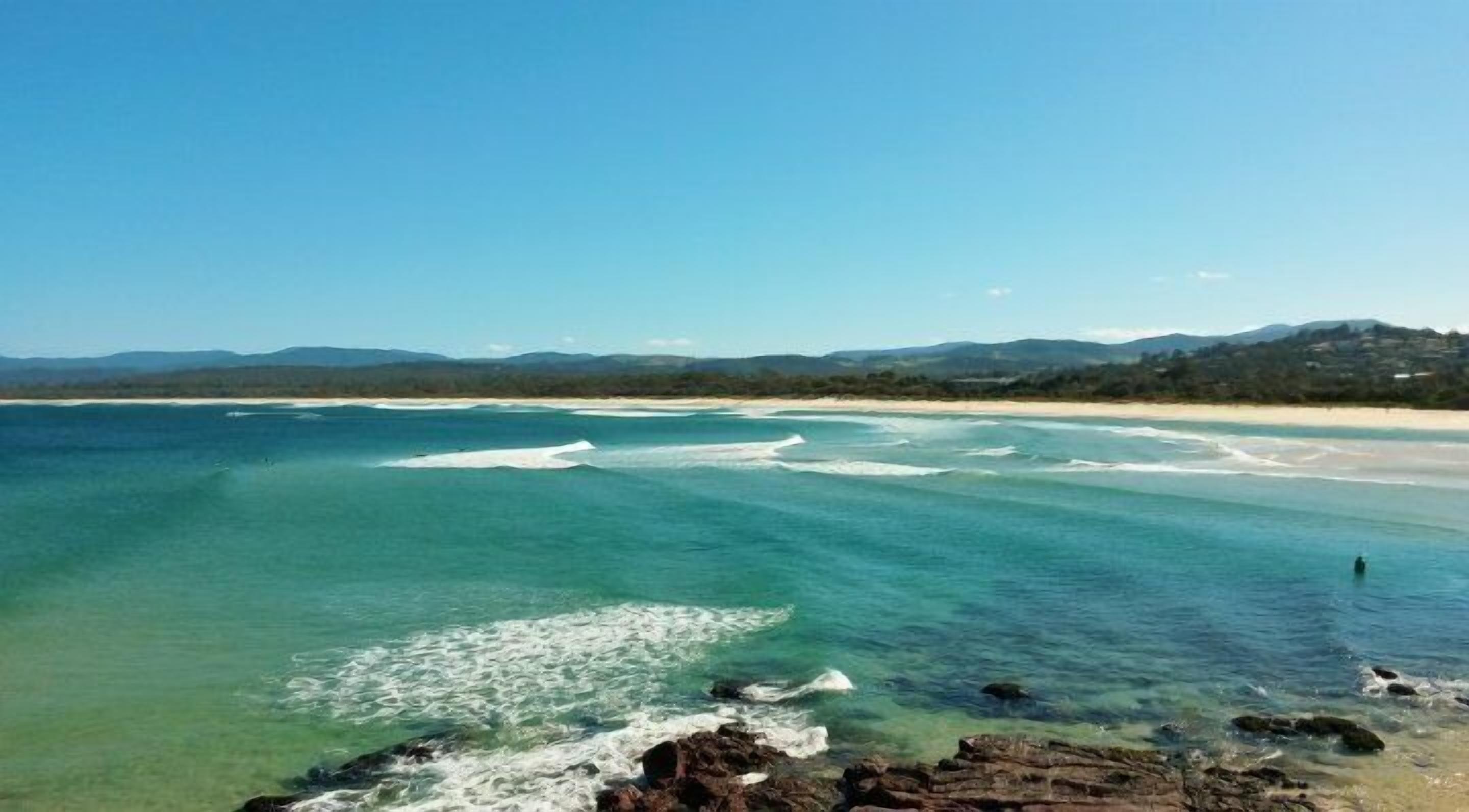 A coastal landscape with clear waters, a sandy beach, and distant hills.