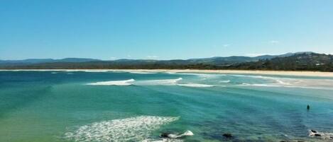 A coastal landscape with clear waters, a sandy beach, and distant hills.