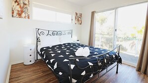 A bedroom with a black and white patterned bedspread, a bedside table with a lamp, and a view of the outdoors through a sliding glass door. - Beachcomber: Unit 3 (Upstairs) (Merimbula)