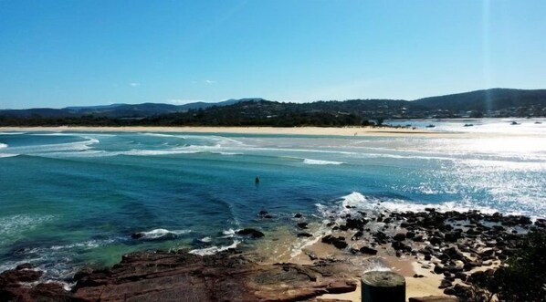 A coastal beach scene with clear blue waters, sandy shores, and rocky outcrops.