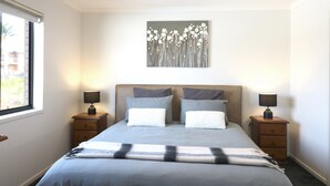 A bedroom with a bed, two nightstands, a window, and a wall-mounted artwork. - Beach Break: Pet friendly fenced yard in town (Merimbula)