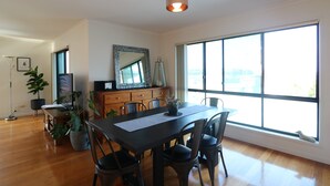 A dining area with a large window, wooden flooring, a table with chairs, a mirror, and a potted plant. - Beach Break: Pet friendly fenced yard in town (Merimbula)