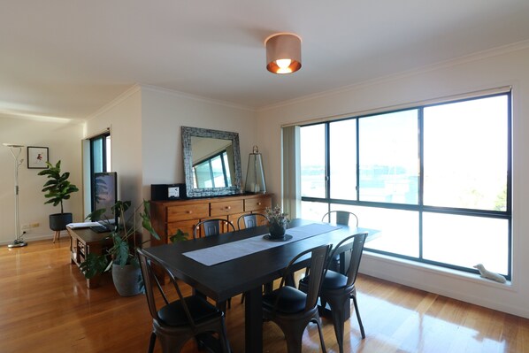 A dining area with a large window, wooden flooring, a table with chairs, a mirror, and a potted plant. - Beach Break: Pet friendly fenced yard in town (Merimbula)