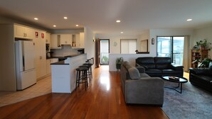A spacious living area with a grey sofa, a black leather couch, and a small coffee table. There is a kitchen with white cabinets and a refrigerator. The room has wooden flooring and a large window providing natural light. - Beach Break: Pet friendly fenced yard in town (Merimbula)