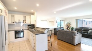 A modern kitchen with white cabinets, a built-in oven, and a bar area. There is a grey sofa and a leather couch in the living area, with a large window providing a view of the outside. - Beach Break: Pet friendly fenced yard in town (Merimbula)