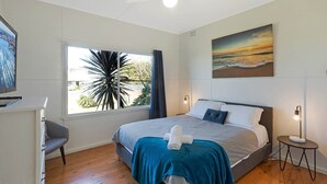 Un dormitorio con cama, escritorio, silla, ventana con cortinas, ventilador de techo, un cuadro y una mesita de noche con lámpara.