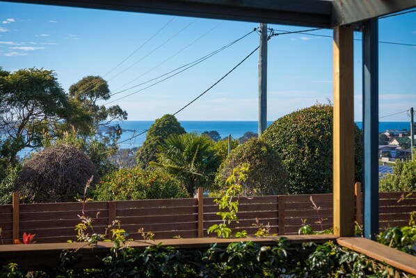 A view from a window overlooking a coastal area with a wooden fence, greenery, and a clear blue sky.