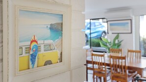 A dining area with wooden chairs and a table, a framed artwork of a van and surfboard, and a car parked outside. - Capri Apartments: 2 bedroom (We have 1 upstairs and one downstairs apartment) (Merimbula)