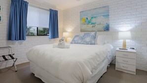 A bedroom with a large bed, white brick walls, a ceiling fan, and a painting of a beach scene. - Capri Apartments: 2 bedroom (We have 1 upstairs and one downstairs apartment) (Merimbula)
