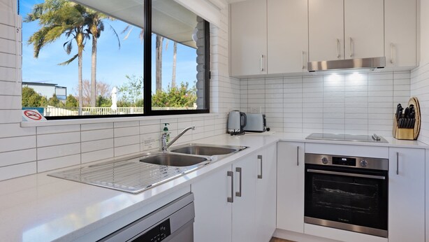 A modern kitchen with white cabinets, stainless steel appliances, a built-in oven, and a window overlooking palm trees. - Capri Apartments: 2 bedroom (We have 1 upstairs and one downstairs apartment) (Merimbula)