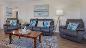 A living room with a white brick wall, two leather sofas with blue floral cushions, a wooden coffee table, and a floor lamp. - Capri Apartments: 2 bedroom (We have 1 upstairs and one downstairs apartment) (Merimbula)