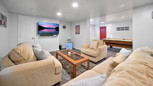 A modern living room with beige sofas, a glass coffee table, a pool table, and a flat-screen TV displaying a scenic landscape. - Poconos Peaceful Retreat. Short drive to Ski, Swim, Hike, or Shop! 4 Bedrooms. (Tobyhanna)