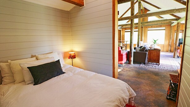 Een slaapkamer met een groot bed, nachtkastje en een lamp. Er is een houten plafond en een deur die leidt naar een balkon.