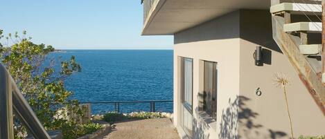 A modern building with a balcony overlooking the sea, a paved pathway, and a number '6' on the wall.