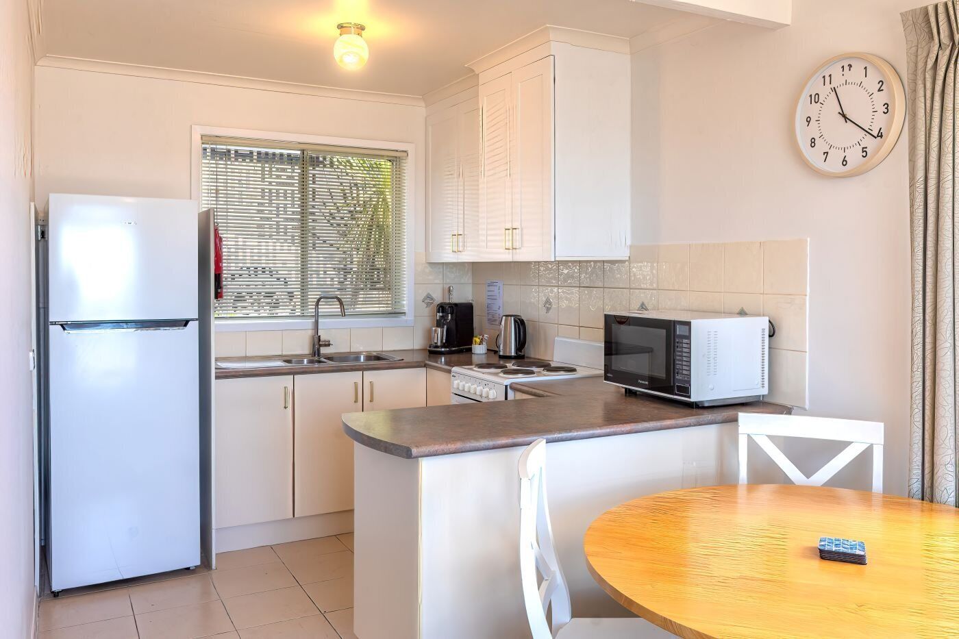 A compact kitchen with a refrigerator, microwave, stove, and sink. There is a dining table with chairs and a clock on the wall.