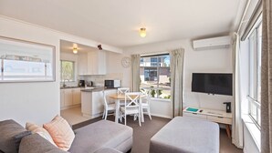 En moderne stue med en grå sofa, skammel og et fladskærms-TV. Der er et spiseområde med et bord og stole, og et køkken i baggrunden.