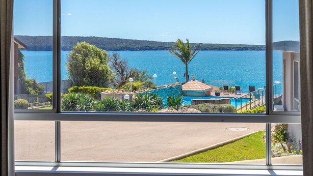 Udsigt fra et vindue over et swimmingpool, en klar blå sø og en fjerntliggende kystlinje.