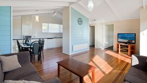 Uma sala de estar moderna com piso de madeira, um sofá, uma mesa de centro e uma TV sobre um suporte de madeira. Há uma área de cozinha com armários brancos e uma mesa de jantar com cadeiras.