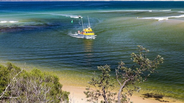 Um barco em um mar azul e límpido, com uma praia de areia e vegetação escassa ao primeiro plano.