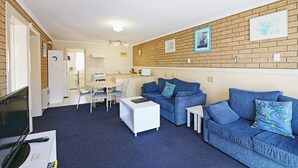 Ein modernes Wohnzimmer mit zwei blauen Sofas, einem weißen Couchtisch und einem Fernseher. Im Hintergrund ist eine Küchenzeile mit einem Esstisch und Stühlen zu sehen.