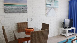 A room with a white brick wall, a dining table with wicker chairs, a TV on a stand, and a blue curtain.