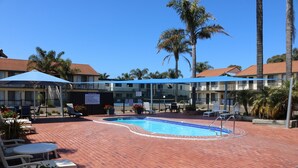 A pool area with lounge chairs, a blue umbrella, and a building with a red roof in the background. - Aquarius Merimbula 3 bedroom Lake View (Merimbula)