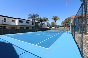 Ein Tennisplatz mit Netz, umgeben von einem Maschendrahtzaun und Palmen.